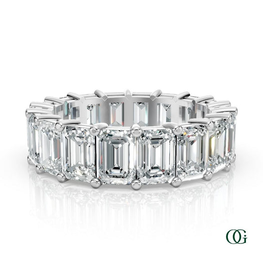 W | A white gold eternity ring featuring a row of emerald-cut clear diamonds, set against a clean white background with a soft reflection.