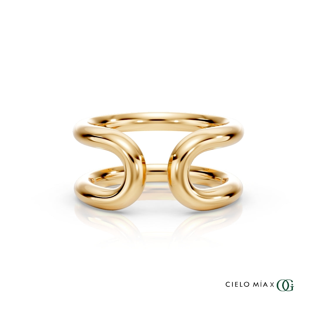 Y| A polished gold double-band ring with a sculptural, intertwined curved design, displayed on a clean white background with a soft reflection and the brand logo 'CIELO MIA x OG' in the bottom right corner.